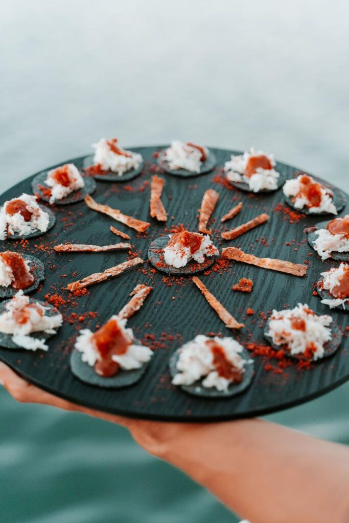 A black round platter with crackers topped with white cream and a reddish garnish, arranged in a circle, with thin strips of food radiating from the center.