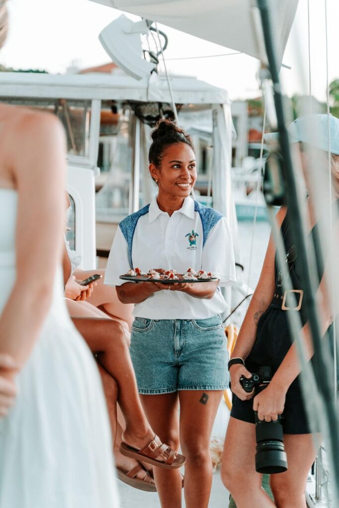 A woman in a white shirt and denim shorts holds a tray of snacks while smiling at a group of people on a boat.