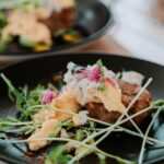 A close-up of a plated dish featuring a patty topped with a creamy yellow sauce, microgreens, and edible flowers, served on a black plate.