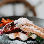 A plated dish featuring a piece of glazed meat on a waffle, garnished with a crisp slice, cream, herbs, and powdered sugar.