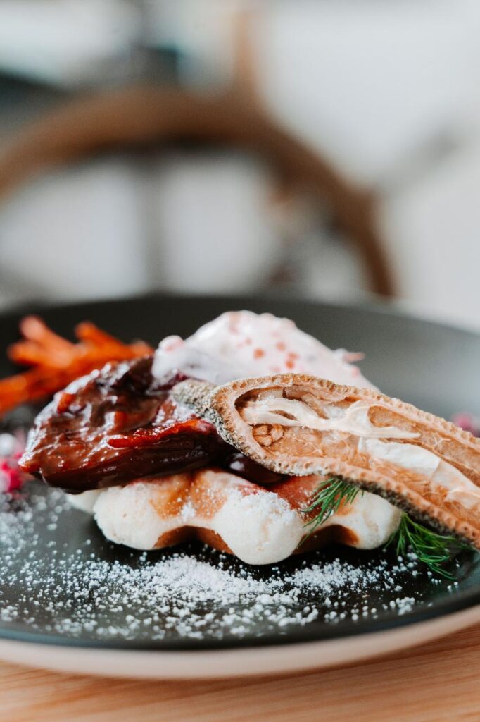 A plated dish featuring a piece of glazed meat on a waffle, garnished with a crisp slice, cream, herbs, and powdered sugar.