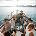 A group of people sit on a boat as a musician plays guitar and sings; the boat is on calm water with buildings visible in the background.