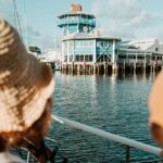 Two people on a boat look toward a blue waterfront building with large windows and a tower, under a partly cloudy sky.