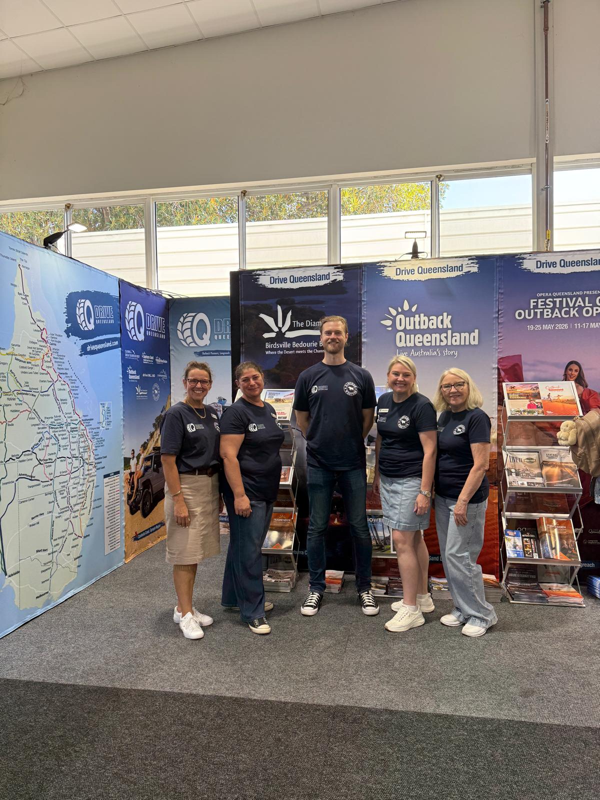 Five people stand smiling in front of tourism display booths for Drive Queensland and Outback Queensland, with brochures and a large map in the background.