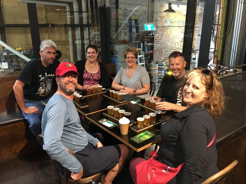 Six people sit around a table with beer flight trays and a coffee cup in an industrial-style brewery or bar setting, smiling at the camera.