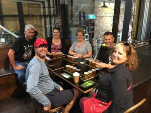 Six people sit around a table with beer flight trays and a coffee cup in an industrial-style brewery or bar setting, smiling at the camera.