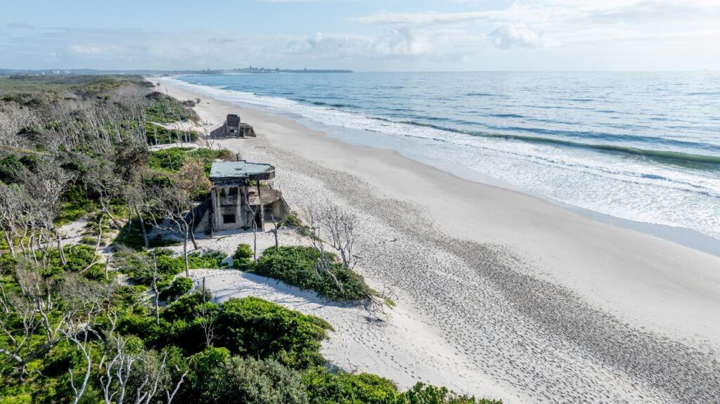 Aerial view of a sandy beach with waves, coastal vegetation, and an old concrete structure near the shoreline.