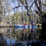 Two people paddle a kayak on a calm, reflective body of water surrounded by tall, leafless trees; another person is visible in a separate kayak in the background.