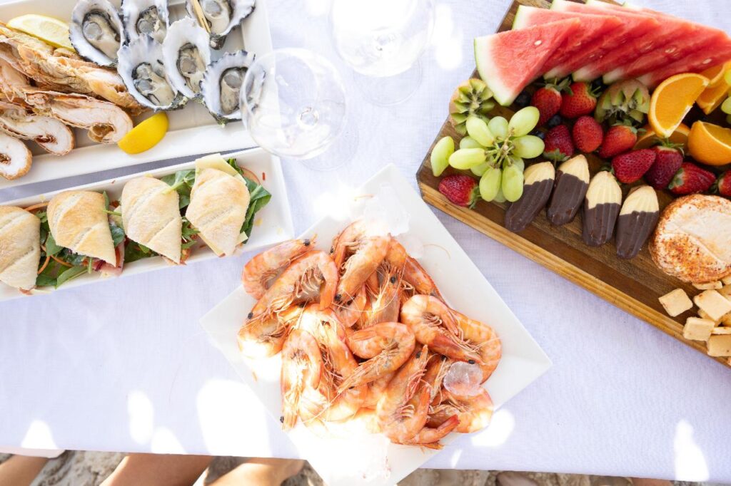 A table set with a platter of shrimp, fresh oysters, sandwiches, chocolate-dipped fruit, assorted cheeses, watermelon, grapes, strawberries, and orange slices on white dishes.