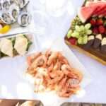 A table set with a platter of shrimp, fresh oysters, sandwiches, chocolate-dipped fruit, assorted cheeses, watermelon, grapes, strawberries, and orange slices on white dishes.