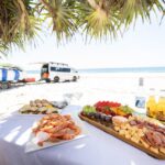 A table with seafood, fruit, and cheese platters is set on a sandy beach, with drinks and a van parked nearby; ocean and surfboards are in the background.