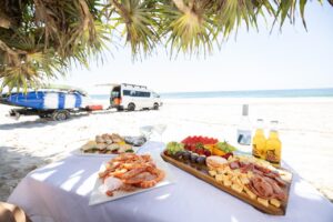 A table with seafood, fruit, and cheese platters is set on a sandy beach, with drinks and a van parked nearby; ocean and surfboards are in the background.