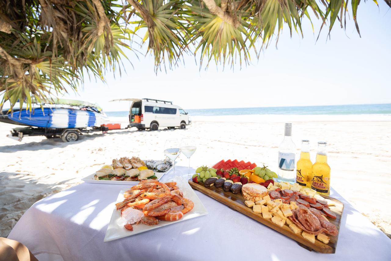 A table with seafood, fruit, and cheese platters is set on a sandy beach, with drinks and a van parked nearby; ocean and surfboards are in the background.