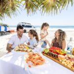 A family sits at a table on a sandy beach, enjoying a seafood and fruit picnic under palm trees, with a van and surfboards in the background near the ocean.