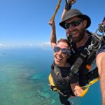 Two people tandem skydiving over clear blue ocean water and reefs, smiling at the camera with a sunny sky above.