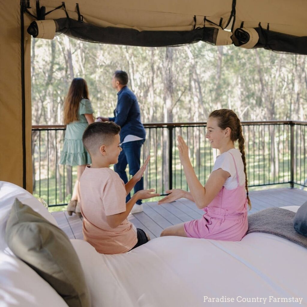 Two children sit on a bed playing a hand clapping game inside a tent, while two adults stand on a balcony overlooking trees outside.