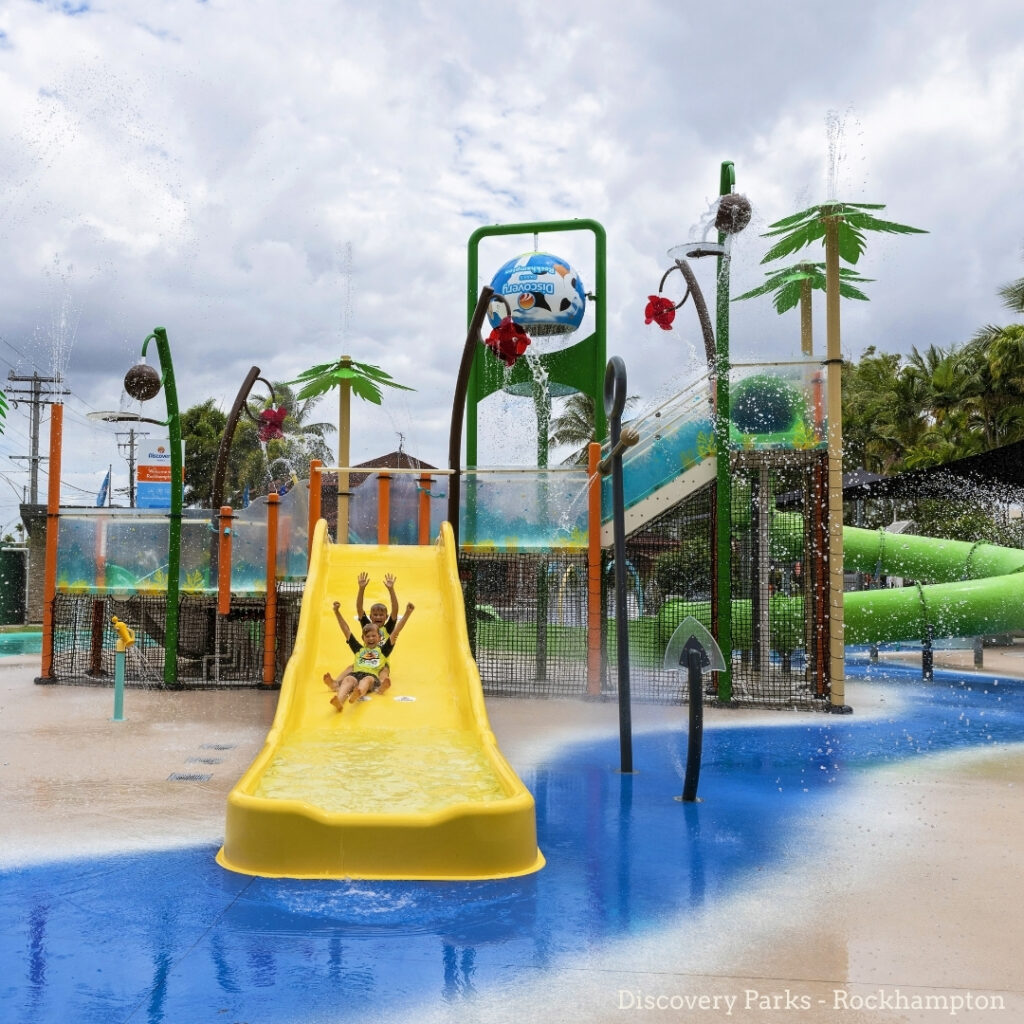 Two children slide down a yellow water slide at an outdoor water play park with palm tree structures and water features.