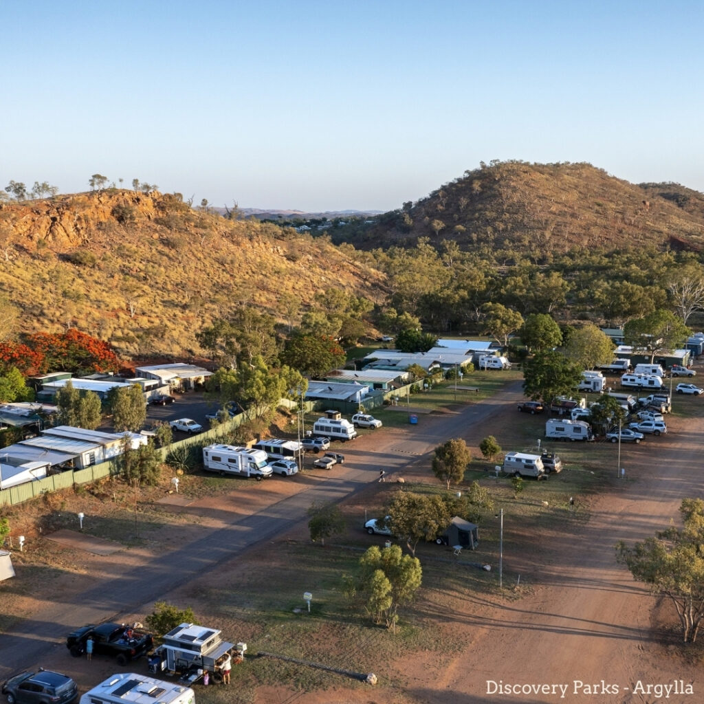 Aerial view of a caravan park with RVs and campers set among trees, surrounded by hills and rocky terrain under a clear sky.
