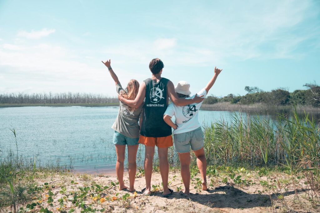 Three people stand with their backs to the camera at the edge of a lake, arms raised and making hand signs, under a blue sky with greenery and water in the background.