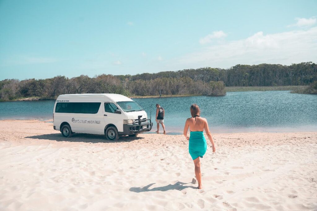 A woman in a turquoise dress walks on sandy beach toward a white van and another person standing near a lake with trees in the background.