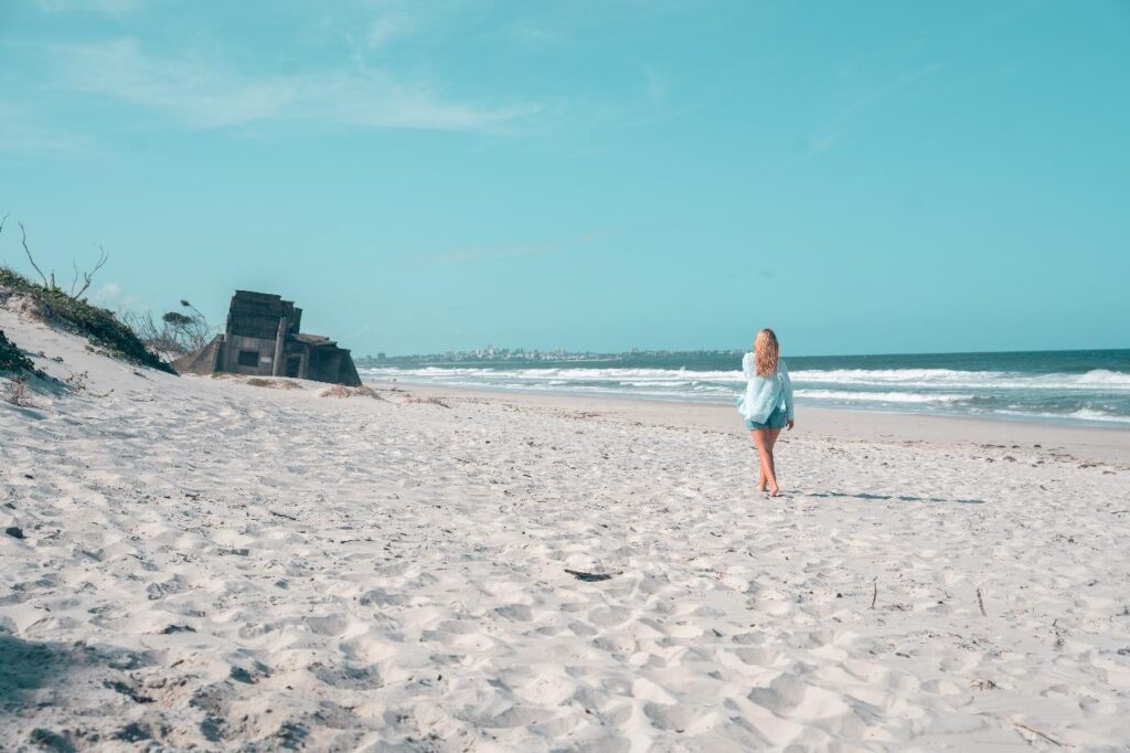 A person walks alone on a sandy beach near the shoreline, with ruins of a concrete structure visible in the distance under a clear blue sky.