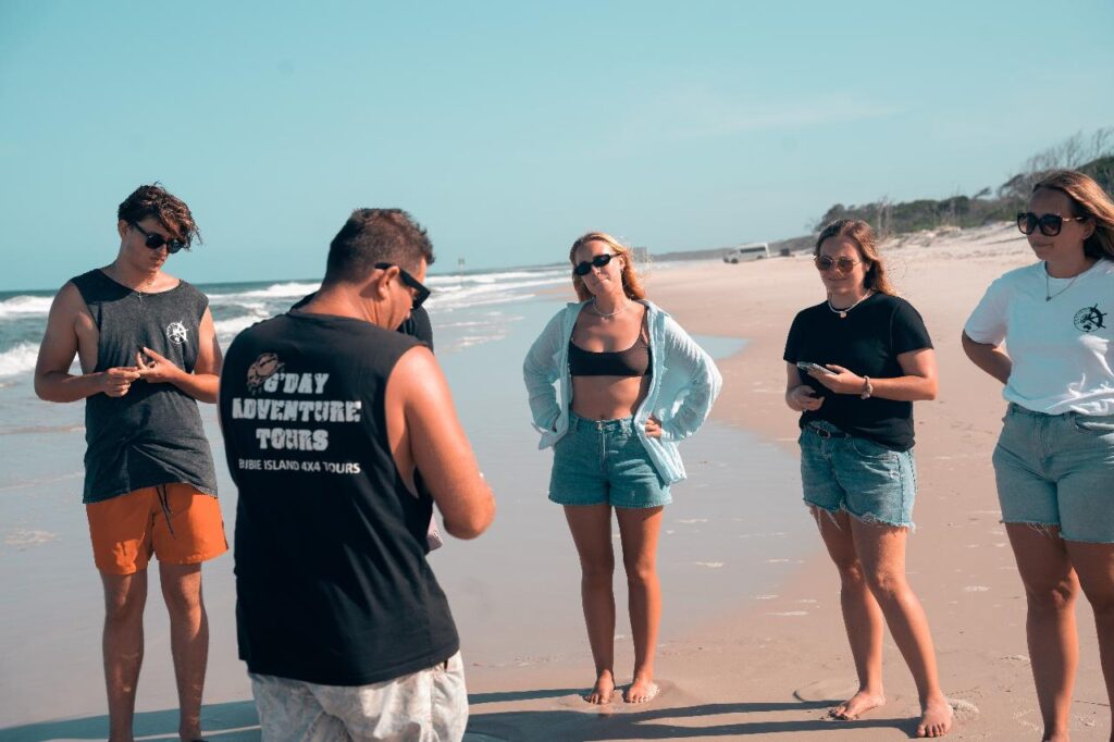 Five people stand on a sandy beach, some looking at their phones and others talking. The sky is clear and the ocean is visible in the background.