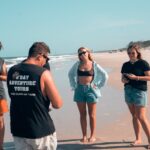 Five people stand on a sandy beach, some looking at their phones and others talking. The sky is clear and the ocean is visible in the background.