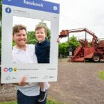 A man holds a young boy while both pose behind a large mock Facebook photo frame; old farming machinery is visible in the background outdoors.