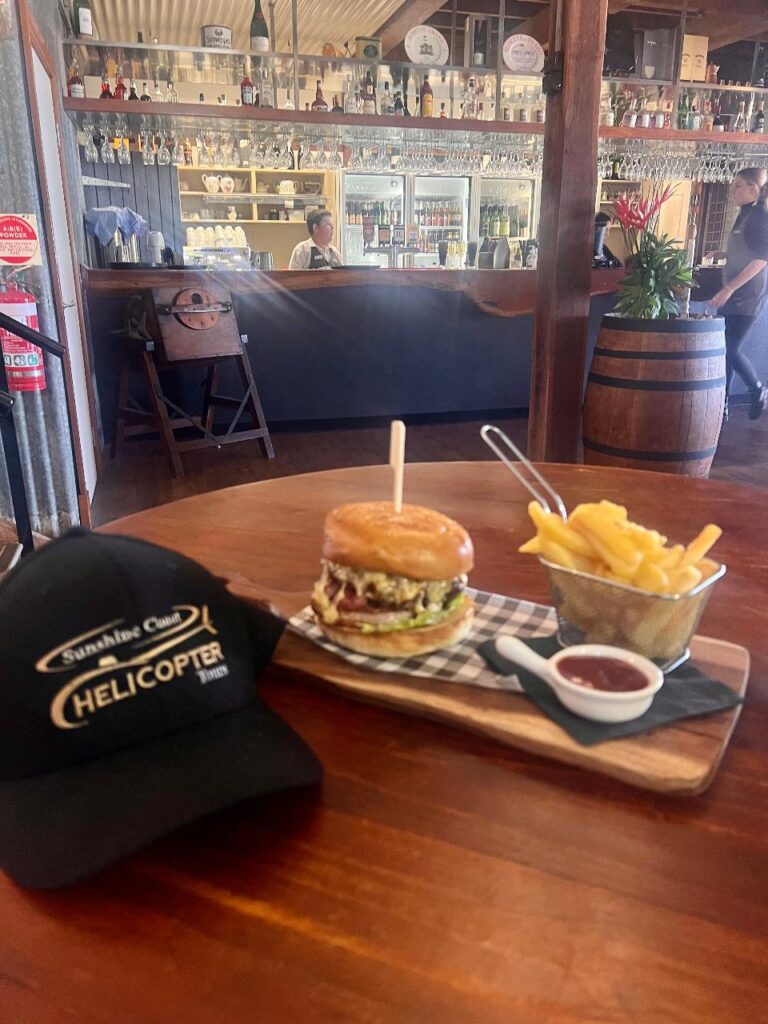 A burger and fries served on a wooden tray with dipping sauce, next to a black cap on a table in a bar-style restaurant.