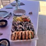 A rectangular plate with oysters, shrimp, lemon wedges, and salmon sits on a white tablecloth, surrounded by dishes, cutlery, and a vase with flowers.