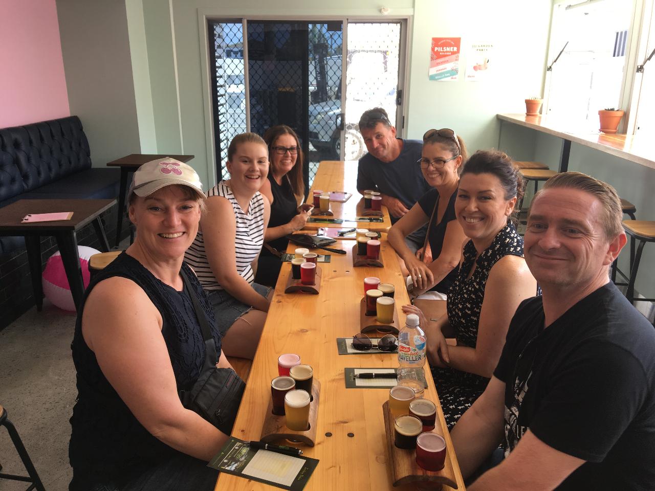 Seven people sit around a wooden table with drinks in a bright indoor space, looking at the camera and smiling.