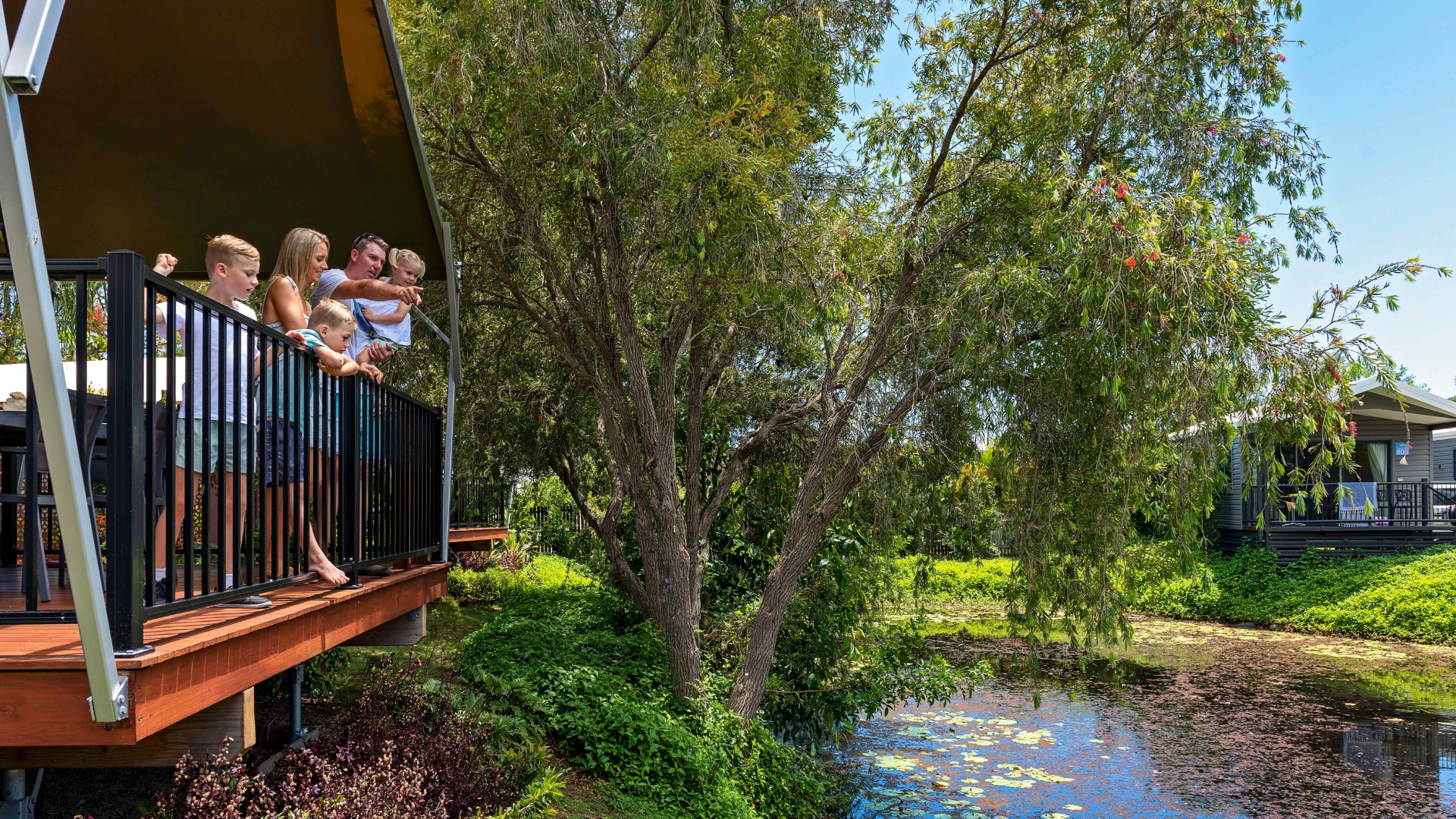 A group of people stand on a balcony overlooking a pond surrounded by trees and greenery on a sunny day.