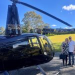 A man and woman stand next to a black helicopter on a sunny day, with a building and trees in the background.