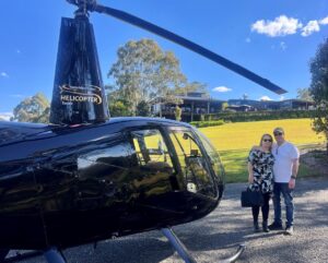 A man and woman stand next to a black helicopter on a sunny day, with a building and trees in the background.