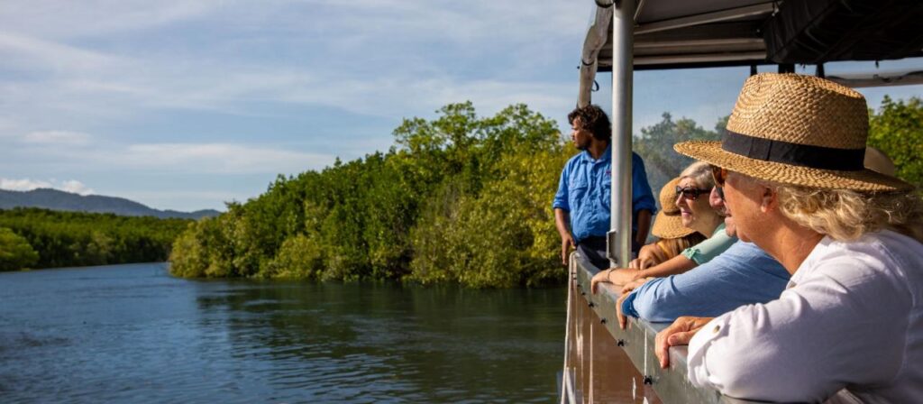 A group of people wearing hats stand on a boat, looking out at a river surrounded by green trees under a blue sky.