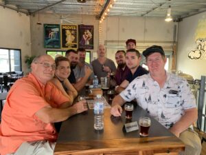 A group of ten people sit together at a table in a brewery or bar, drinks and water bottles on the table, with posters and brewing equipment visible in the background.