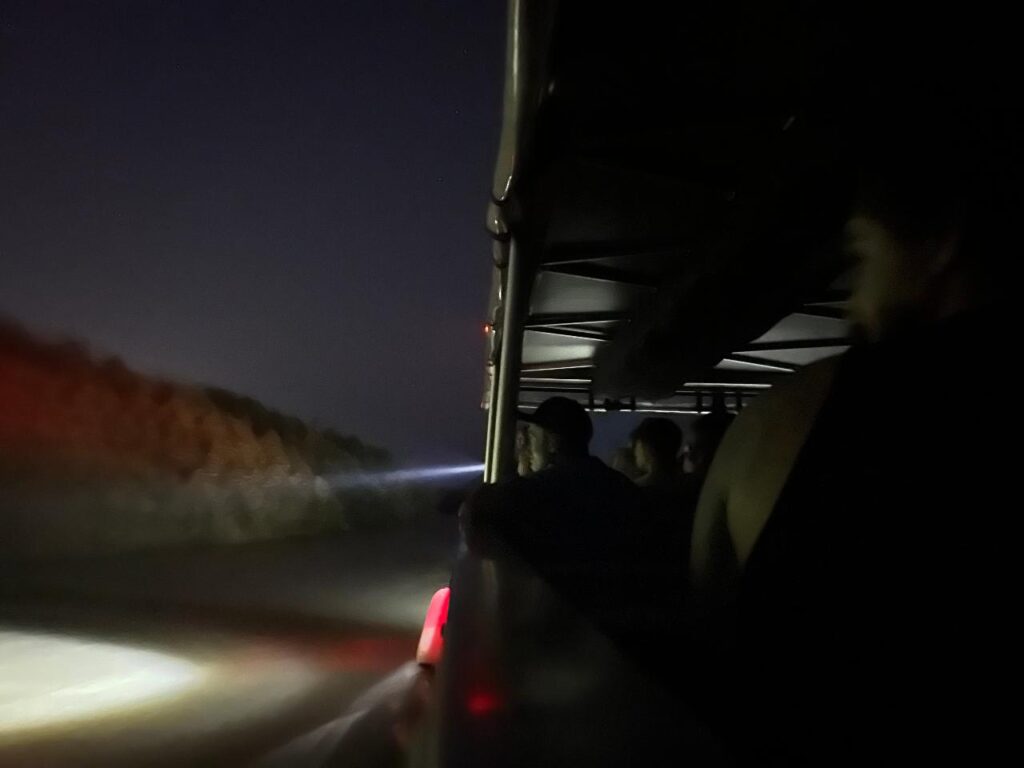 People ride in an open vehicle at night, looking out into the dark as headlights and a beam of light illuminate the road ahead.