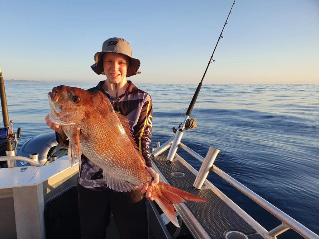Person standing on a boat holds a large fish with both hands; fishing rod and calm water are visible in the background.