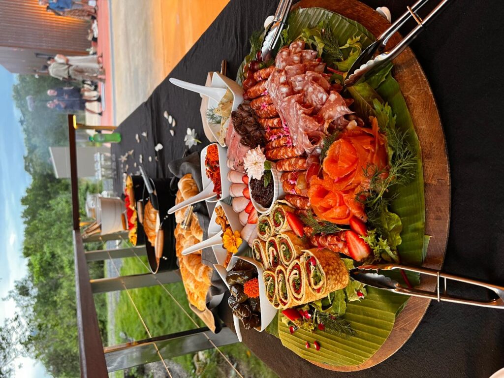 A large platter of assorted cold cuts, shrimp, smoked salmon, and rolled appetizers sits on a black tablecloth at an outdoor buffet.