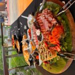 A large platter of assorted cold cuts, shrimp, smoked salmon, and rolled appetizers sits on a black tablecloth at an outdoor buffet.