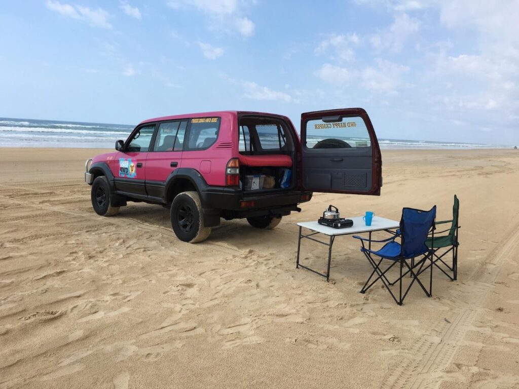 A pink SUV with its rear door open is parked on a sandy beach next to a folding table, two chairs, and camping items. The ocean and sky are in the background.