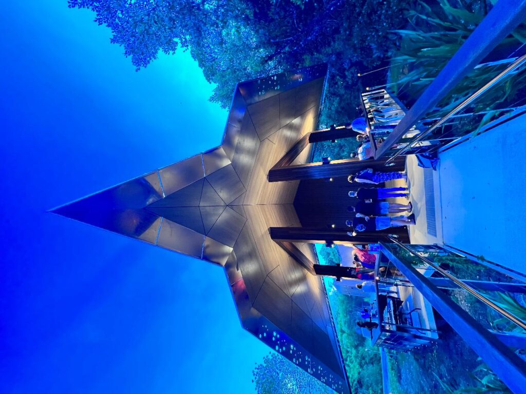 A group of people stands beneath a modern, angular metal pavilion at dusk, surrounded by trees and illuminated by blue lighting.