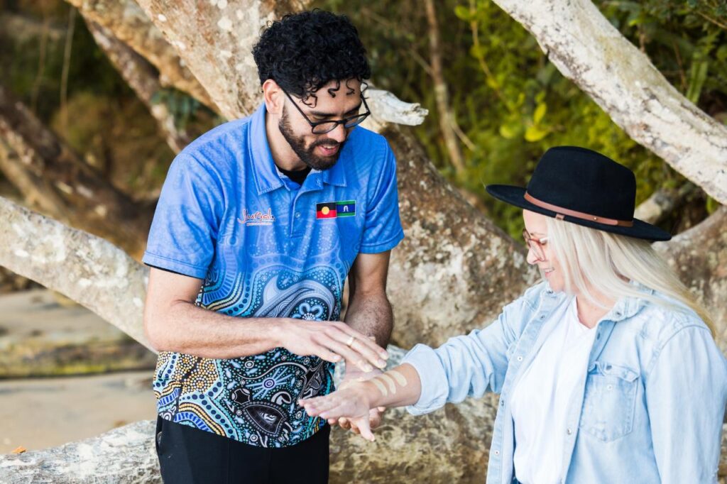 A man in a blue patterned shirt applies ochre paint to a woman’s forearm outdoors, with large tree branches in the background.