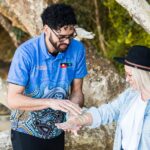A man in a blue patterned shirt applies ochre paint to a woman’s forearm outdoors, with large tree branches in the background.