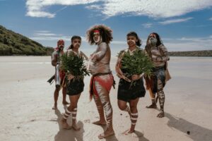 Five people in traditional attire with body paint stand on a sandy beach holding green plants, with blue sky and greenery in the background.