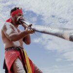 A person with body paint and traditional attire plays a didgeridoo outdoors under a partly cloudy sky.