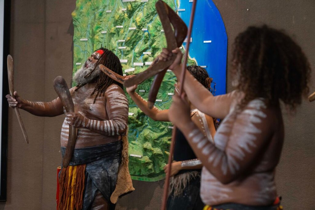 Three people with traditional body paint and attire hold boomerangs, performing in front of a wall decorated with green and blue elements.