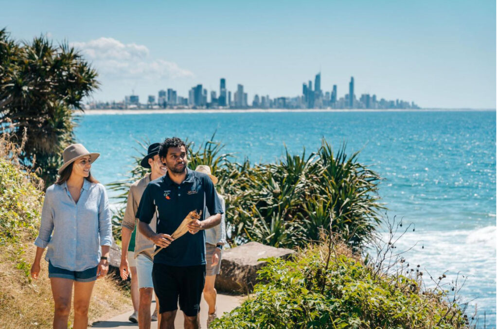 A group of people walk along a coastal path with city skyscrapers in the background and the ocean to their right on a sunny day.