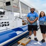 Two people in matching blue uniforms stand on a dock next to a white cruise boat with the word "CRUISES" partially visible on the side.