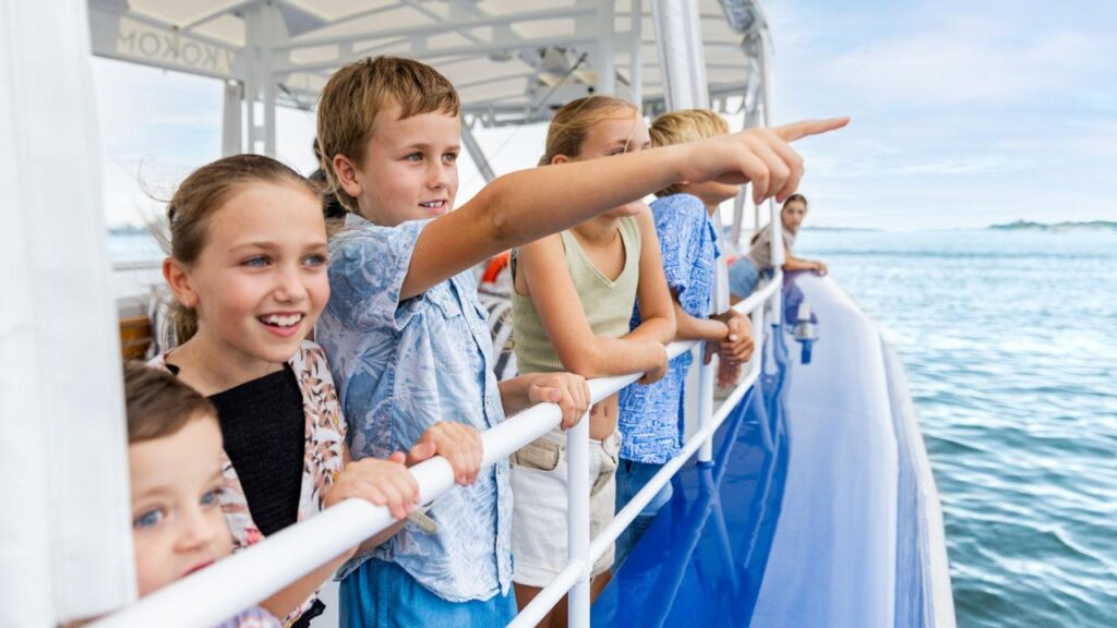 A group of children stand on a boat deck, looking and pointing out at the water, with the sea and sky visible in the background.